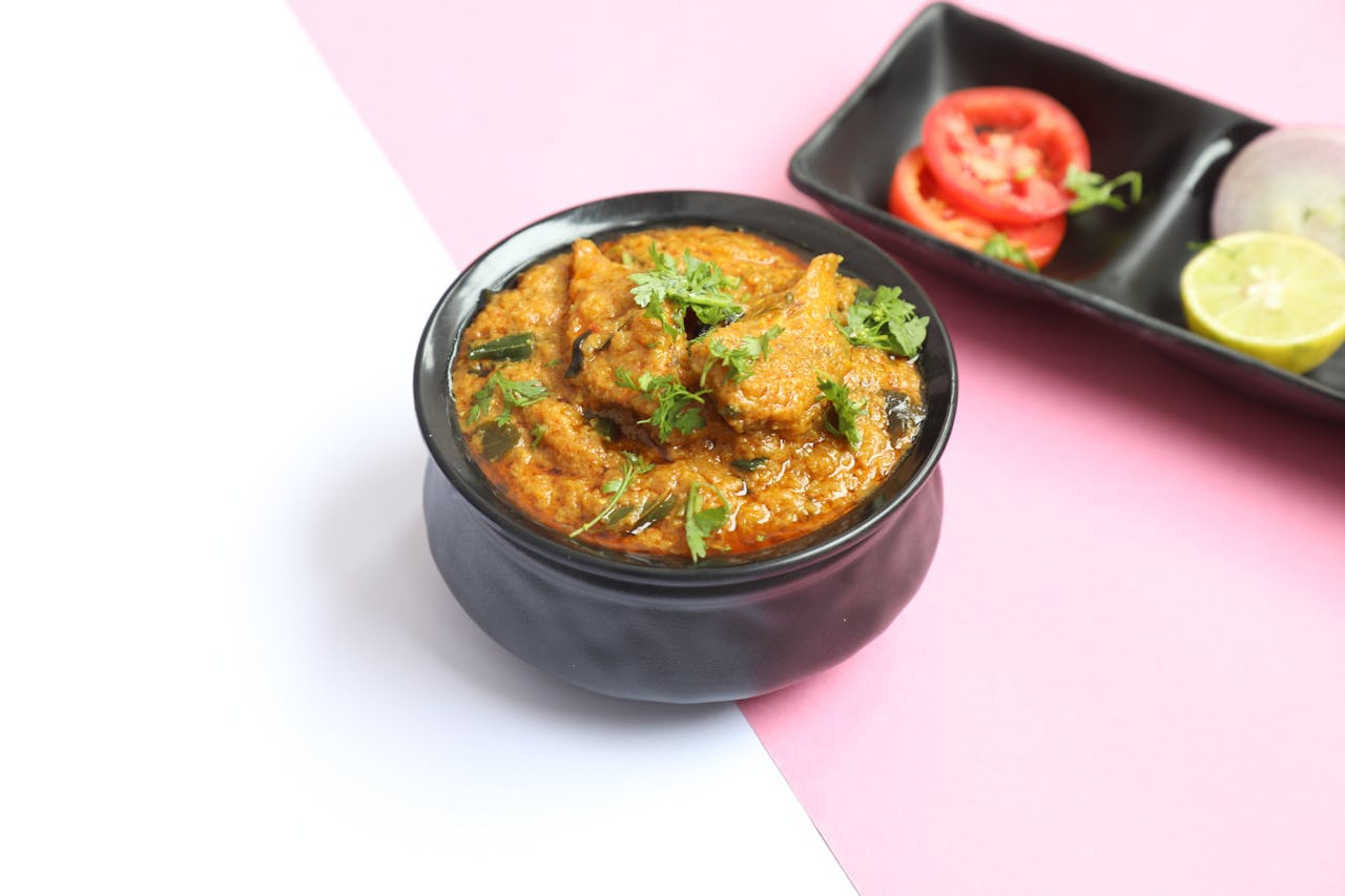 services-01 Spicy Indian curry served in a black bowl with fresh cilantro garnish, accompanied by sliced tomatoes and lemon.