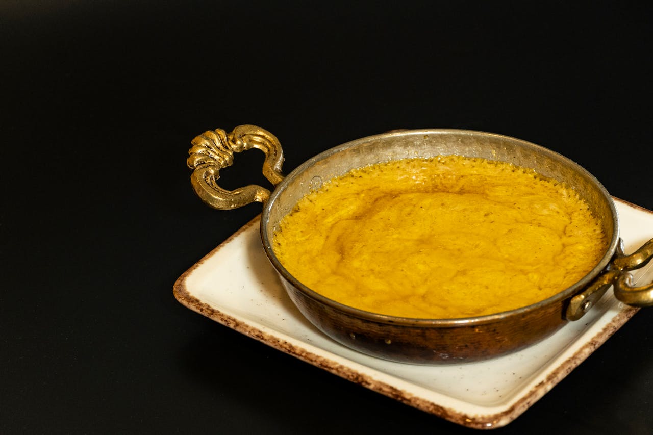 hero-img-01 A traditional Indian dish served hot in a brass kadai on a square plate, against a dark background.